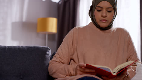 Muslimische-Frau-Mit-Hijab-Sitzt-Zu-Hause-Auf-Dem-Sofa-Und-Liest-Oder-Studiert-Den-Koran